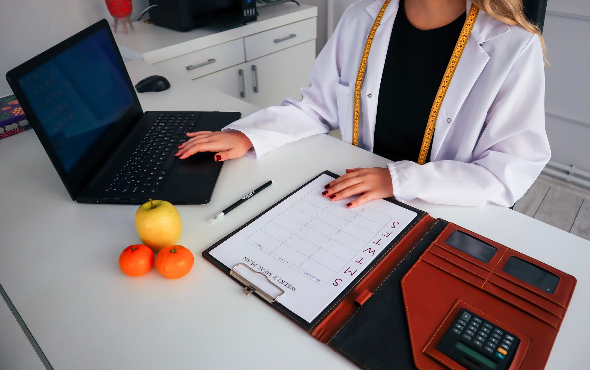 Nutricionista trabalhando em plano de refeições com laptop, frutas e calendário para consulta de saúde.