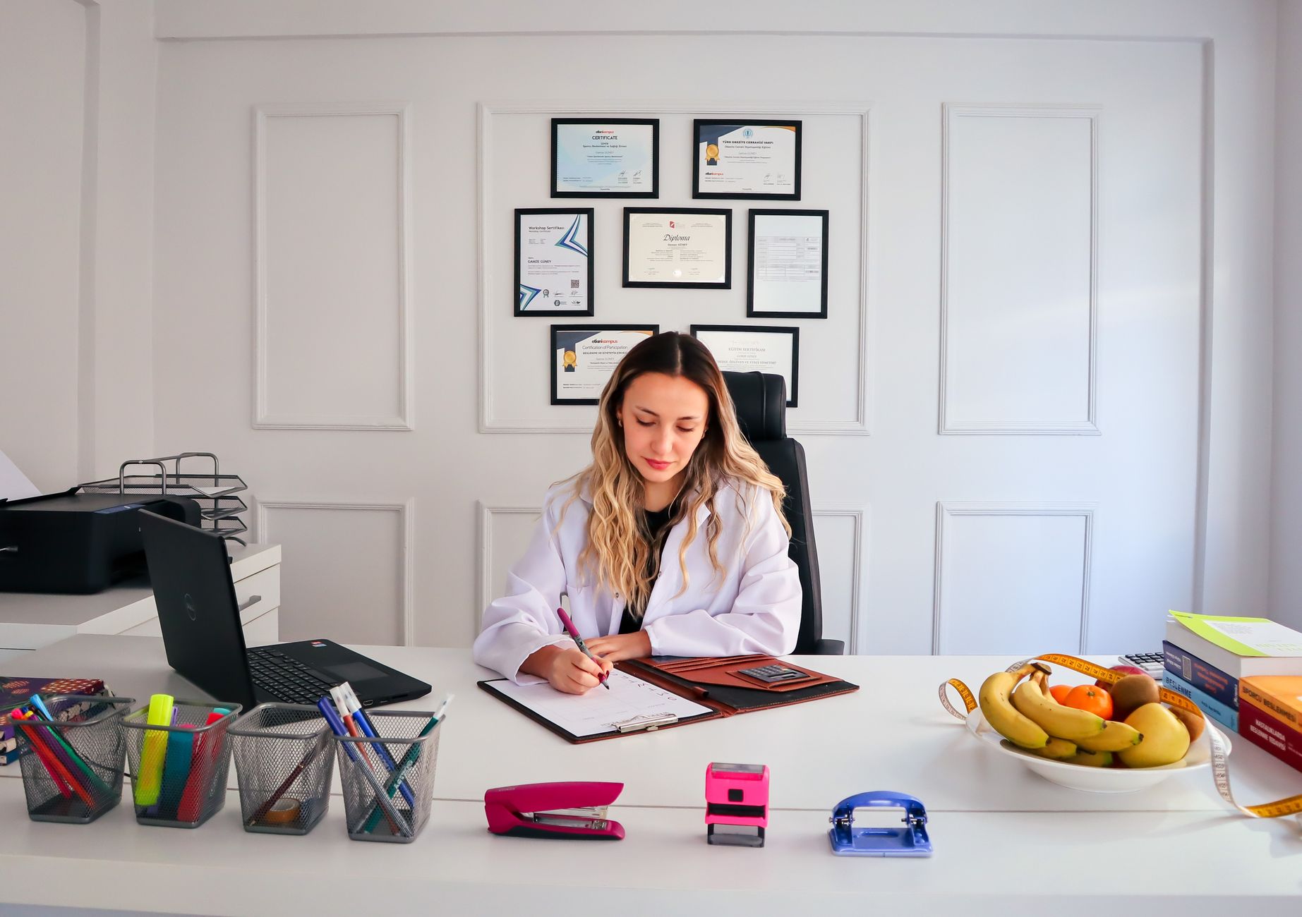 Nutricionista sentado em uma mesa de escritório moderna escrevendo, com frutas e diplomas expostos, enfatizando saúde e profissionalismo.